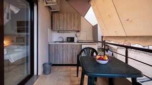 A balcony with a table, a bowl of fruit, a small kitchen area with cabinets and a microwave, and a view of a building through a glass door. - Balcony Studio • Outdoor Kitchen • Uji i Ftohtë (Vlora)