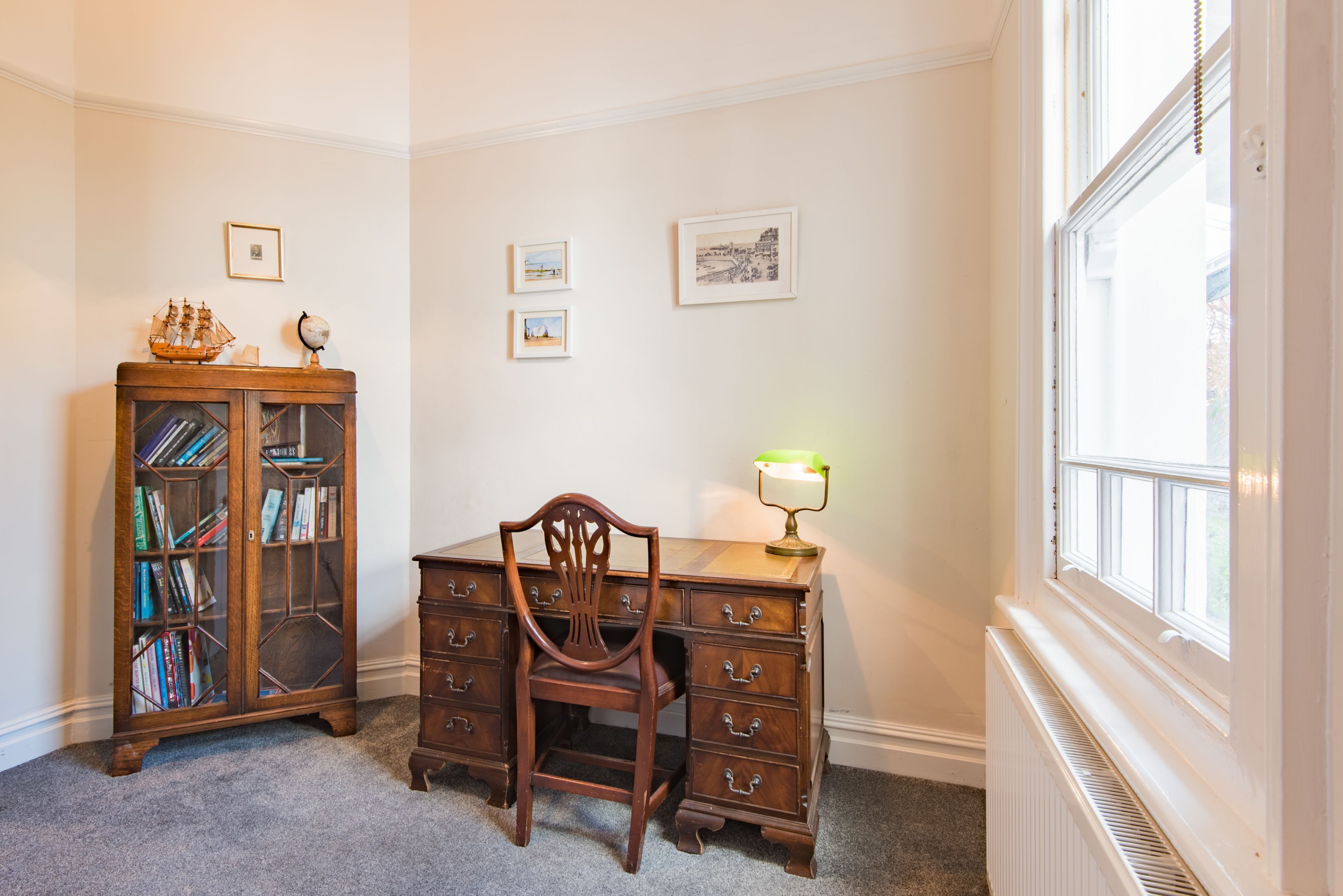 A study room with a wooden desk, a chair, a bookshelf, and framed pictures on the wall.