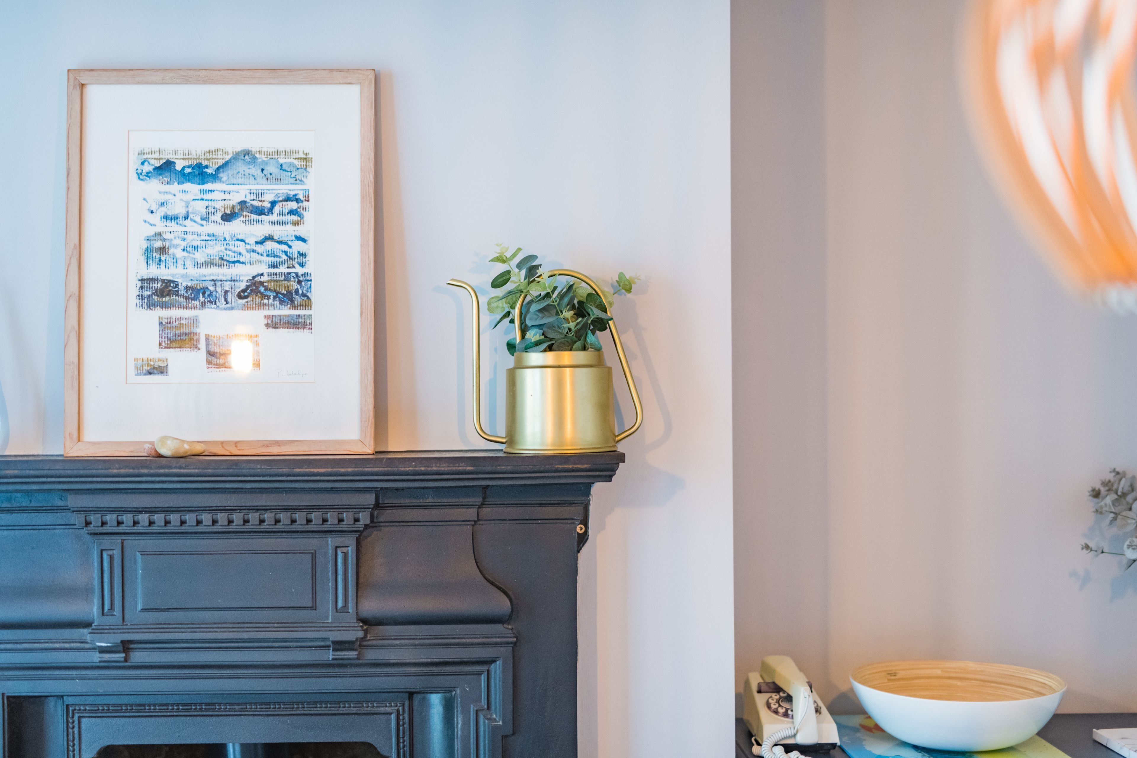 A fireplace with a framed artwork above it, a gold planter with a plant, and a wooden bowl on a table beside the fireplace.
