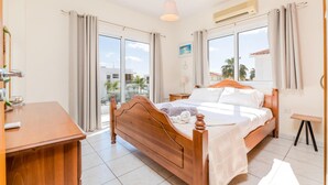 A well-lit bedroom with a wooden bed, a desk, and a view of palm trees through the windows.