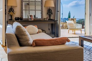 A cozy living room with a sofa, pillows, and a coffee table. There is a balcony with a chair and a view of the outdoors. - Mas Gauda – Garden & family space, Côte d’Azur (La Gaude)