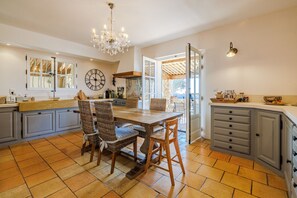 A spacious kitchen with a wooden table and wicker chairs, a chandelier, and a large clock on the wall. - Mas Gauda – Garden & family space, Côte d’Azur (La Gaude)