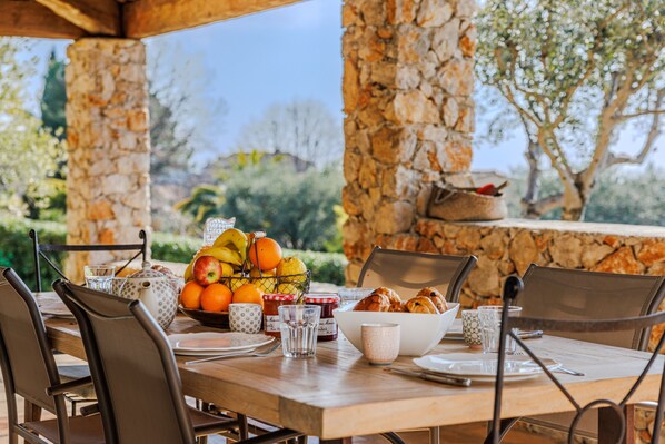 Una mesa de comedor lista para el desayuno, con frutas, pasteles y bebidas, en un entorno al aire libre con pilares de piedra y vistas a zonas verdes.