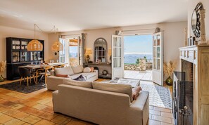 A spacious living room with a large sofa, dining area, and a view of the countryside through open doors. - Mas Gauda – Garden & family space, Côte d’Azur (La Gaude)