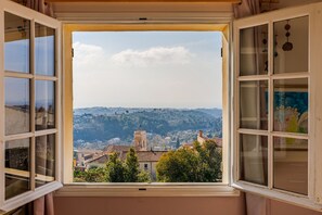 A room with a large window offering a view of a town with buildings and hills. - Mas Gauda – Garden & family space, Côte d’Azur (La Gaude)