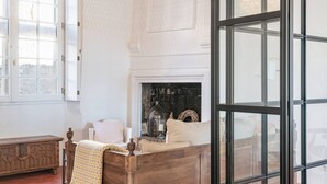 A room with a wooden bed, a white upholstered chair, a small table with a candle, and a fireplace. - Apt Chagall – In the heart of Saint-Paul de Vence (Saint Paul de Vence)