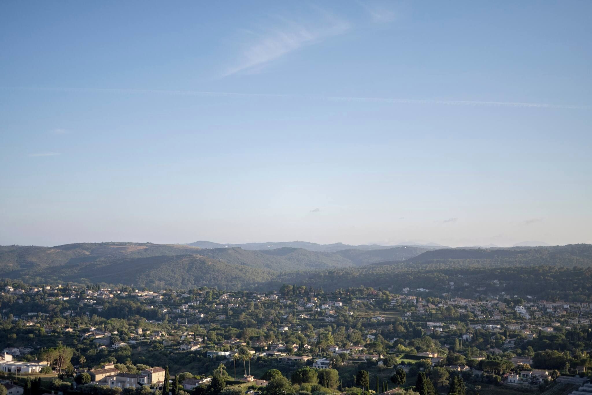 A panoramic view of a town with a mix of residential buildings and greenery, surrounded by hills.