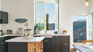 A modern kitchen with dark cabinets, a white island, and a large window offering a view of the countryside. - Apt Chagall – In the heart of Saint-Paul de Vence (Saint Paul de Vence)