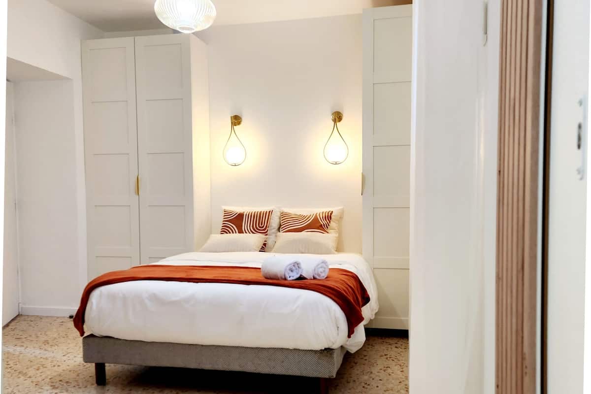 Un lit bien fait, recouvert d’une couvre-lit blanche et marron, deux appliques murales et une grande armoire.