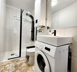 A modern bathroom with a glass shower enclosure, a toilet, a washing machine, and a sink with a mirror above. - Le 56 entre ville&lac sejouraixlesbains (Aix-les-Bains)
