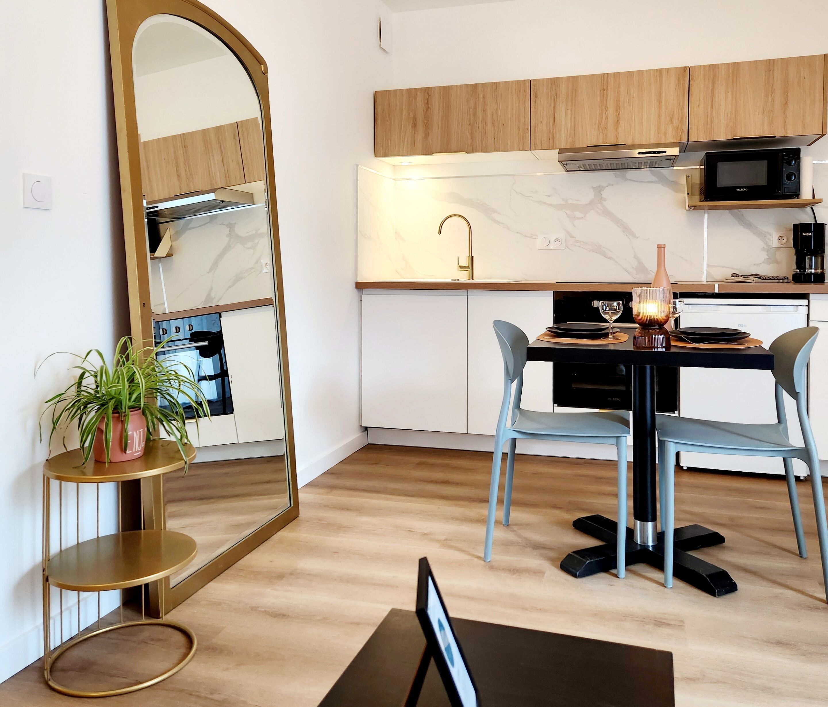 Una cocina moderna con encimera de mármol, armarios de madera, microondas y un lavabo. Hay una mesa de comedor con dos sillas y una planta en maceta.