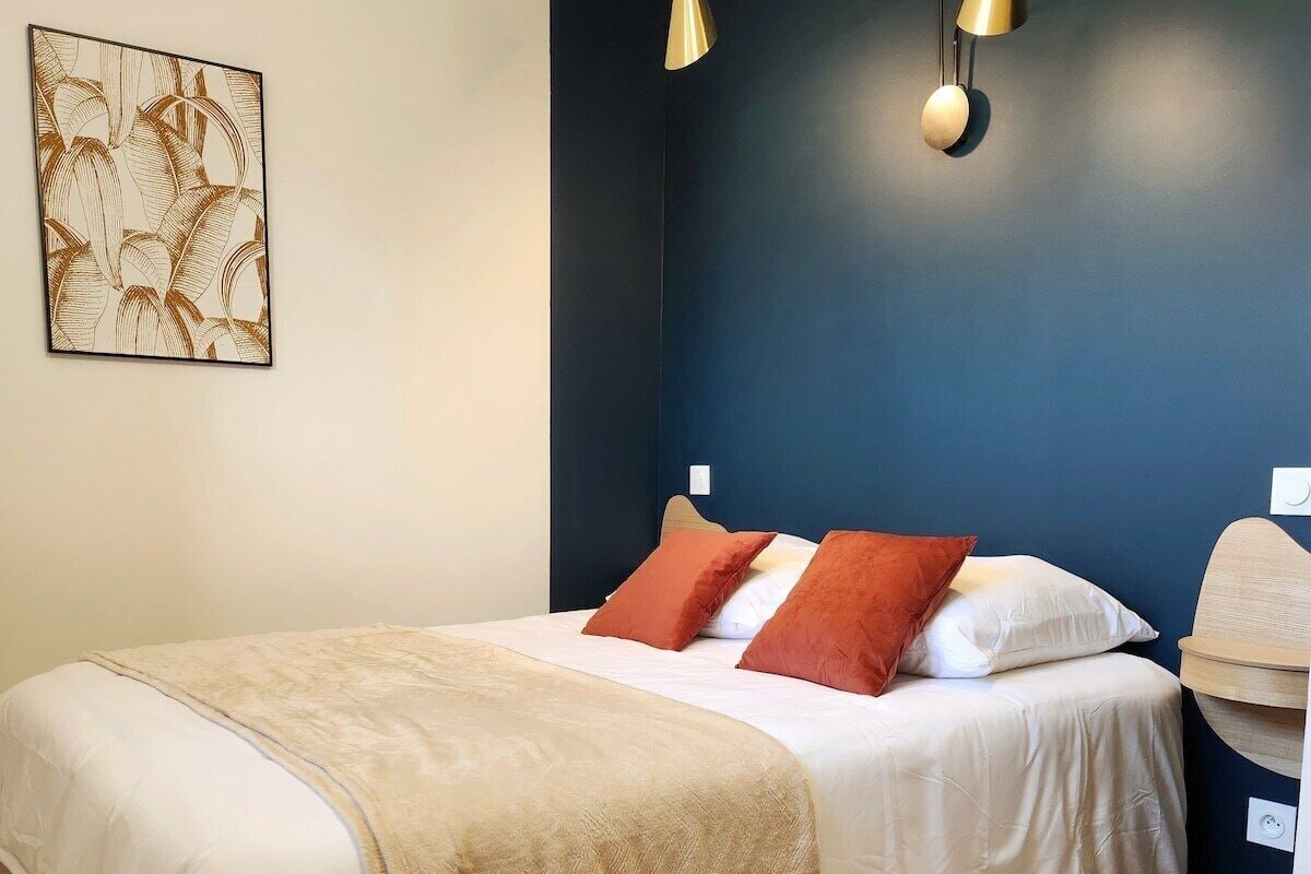 Un lit bien fait, recouvert d’une couvre-lit beige et agrémenté de deux coussins rouges, un mur d’accent bleu foncé et deux lampes fixées au mur.