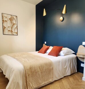 A neatly made bed with a beige bedspread and two red pillows, a dark blue accent wall, and two wall-mounted lamps. - Le 56 entre ville&lac sejouraixlesbains (Aix-les-Bains)