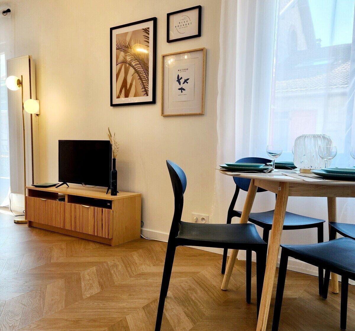 A modern living room with a flat-screen TV on a wooden TV stand, a dining table with chairs, and framed artwork on the wall.