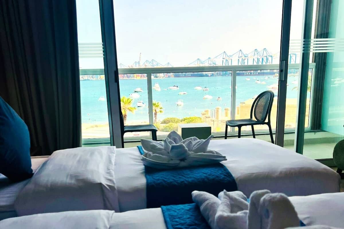 A hotel room with two beds, a view of the sea, and a balcony with a chair.