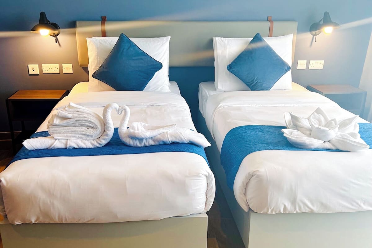 A hotel room with two beds, each with white bedding and blue pillows, and a blue headboard. There are two bedside lamps, a wooden nightstand, and a folded towel on each bed.