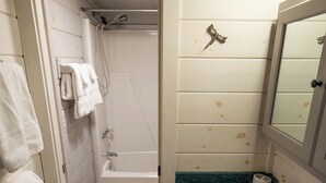 Et badeværelse med en hvid vask, et spejl og en bruser. Der hænger håndklæder på væggen og en flaske sæbe på bordet.