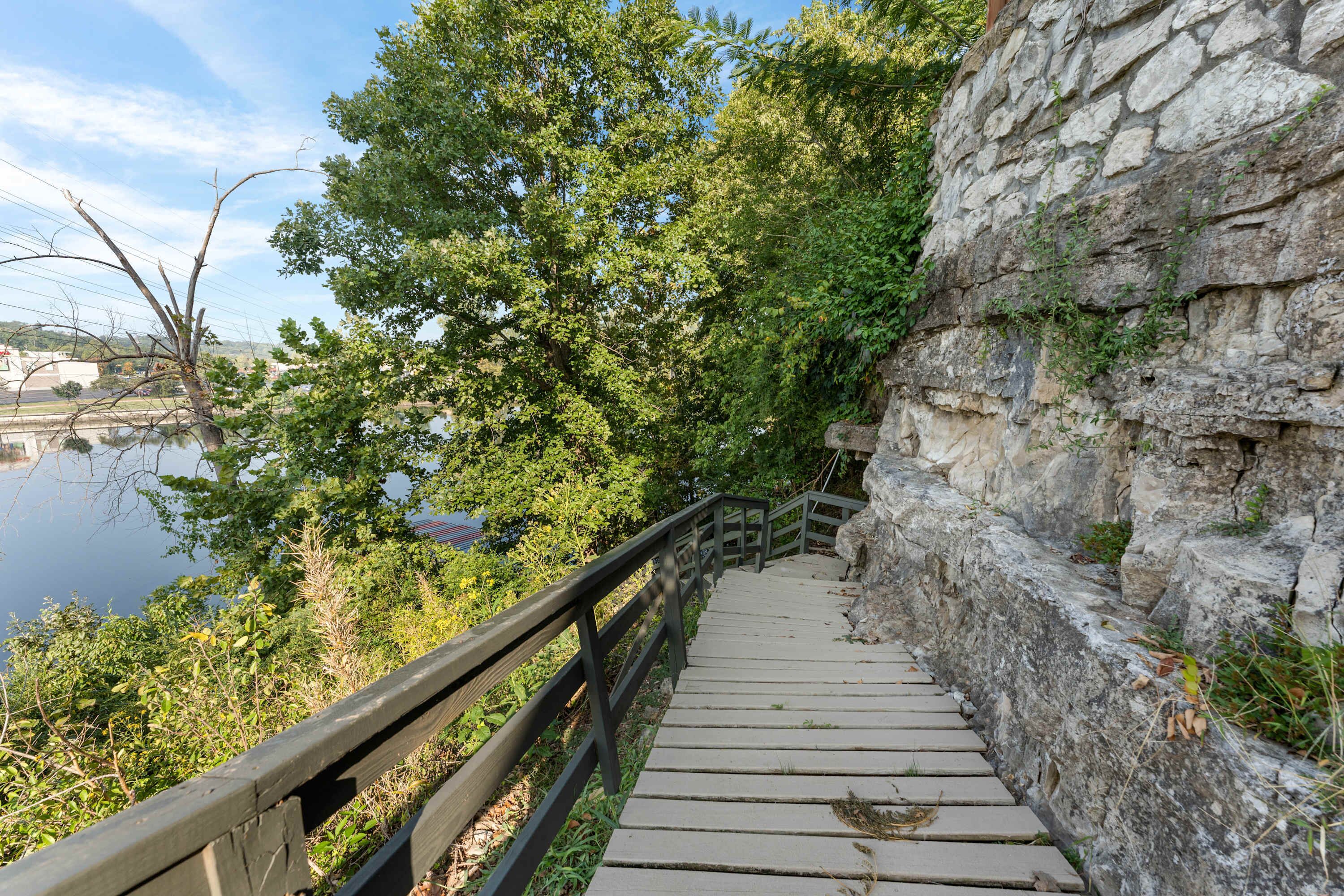 Eine Steintreppe führt zu einem hölzernen Deck mit Geländer, umgeben von viel Grün und mit Blick auf ein Gewässer.