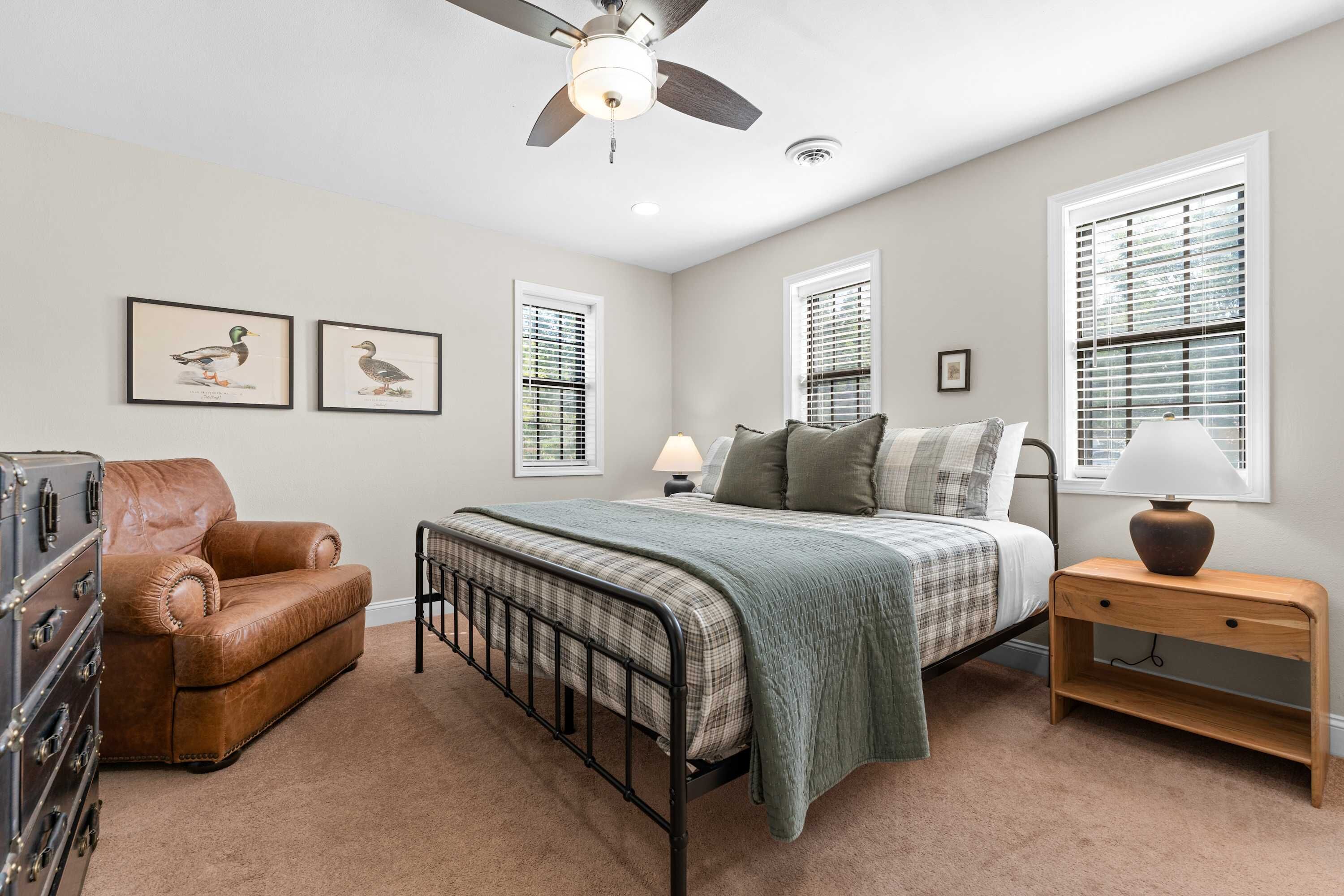 A bedroom with a bed, a dresser, a chair, a nightstand, and two windows with blinds.