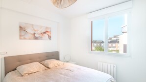 A bedroom with a large bed, a bedside table, a wall-mounted artwork, and a window with a view of buildings.