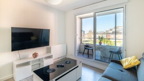 A modern living room with a flat-screen TV, a glass-top coffee table, a sofa, and a sliding glass door leading to a balcony.