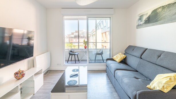 A modern living room with a grey sofa, a flat-screen TV, a glass coffee table, and a balcony with chairs.