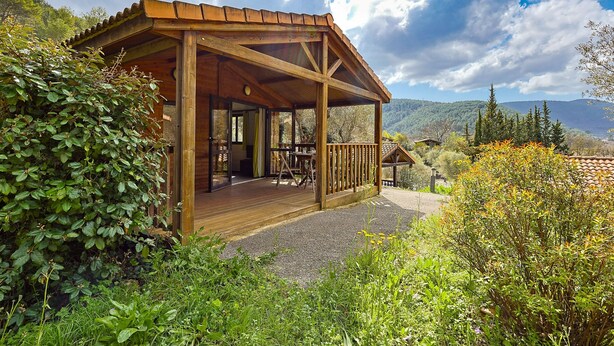 Une cabane en bois dotée d’un porche abrité, entourée d’une végétation luxuriante et avec des montagnes en toile de fond.