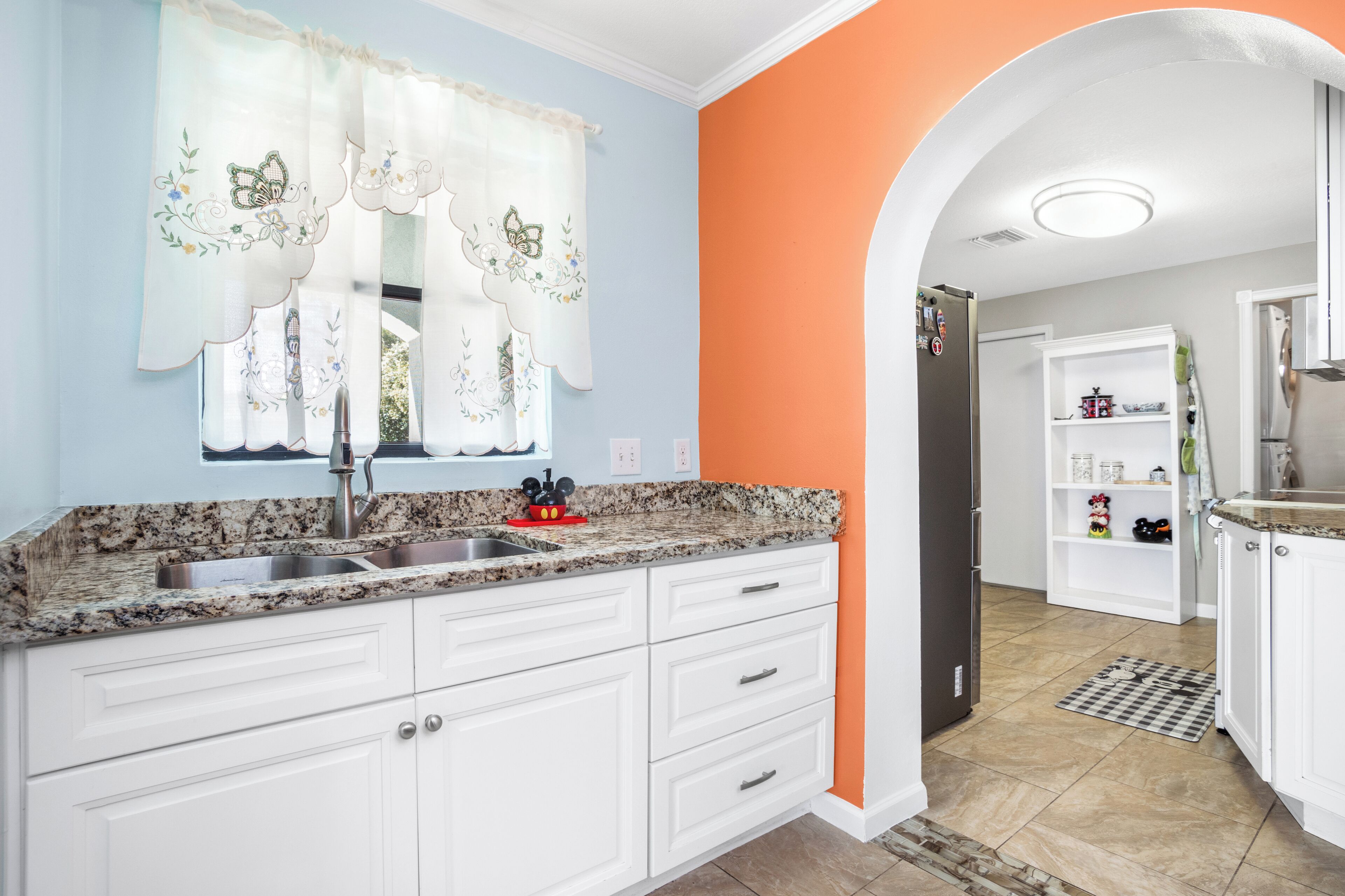 Una cocina con armarios blancos, encimera de granito, pileta de acero inoxidable y una pared de acento naranja.