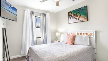 Un dormitorio con cama, cortinas, ventilador de techo, un cuadro de playa y televisor en la pared.