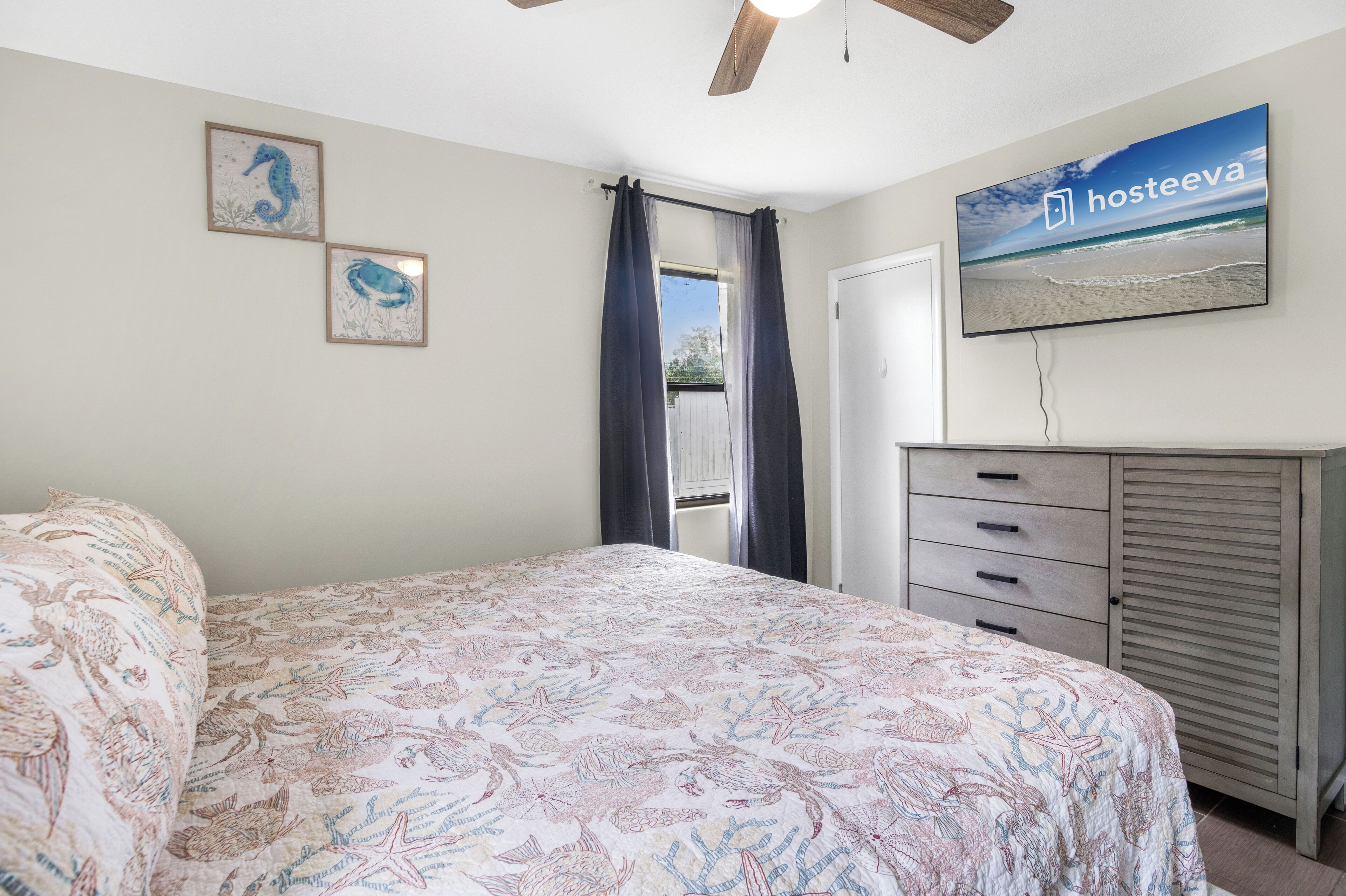A bedroom with a bed, a dresser, a ceiling fan, and a wall-mounted TV displaying 'hosteeva'.