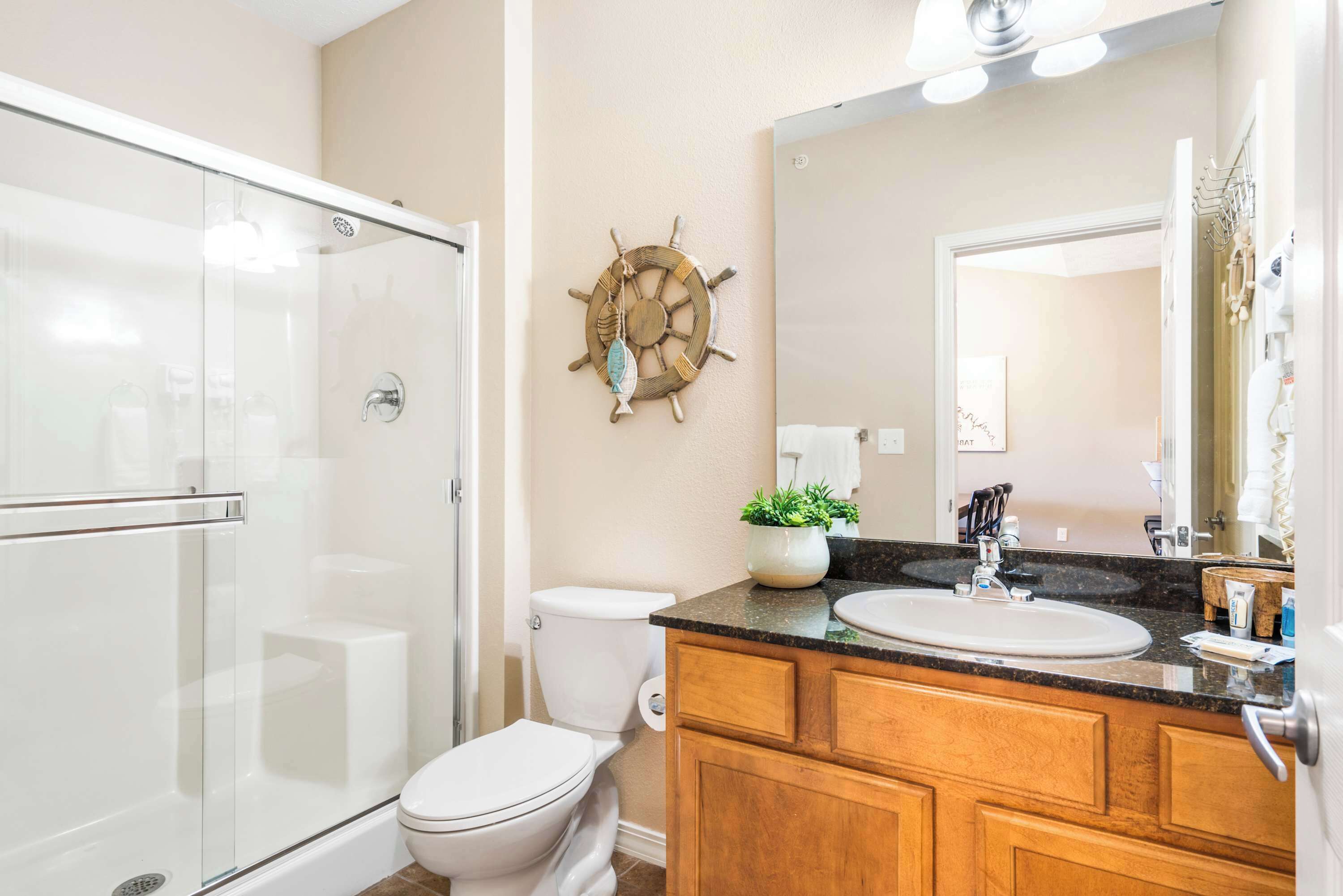 A bathroom with a glass shower enclosure, a toilet, a sink with a vanity, and a decorative ship wheel on the wall.