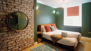 Un dormitorio con pared de ladrillo, un espejo redondo, una cama con varias almohadas, una ventana con persianas y un radiador.