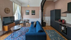 Una sala de estar moderna con un sofá azul, una mesa de centro redonda y un televisor. Se observa también un área de cocina con estufa y armarios.