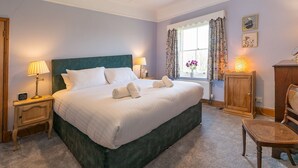 A neatly made bed with white linens, a green headboard, and a wooden nightstand with a lamp. There is a window with floral curtains, a wooden cabinet, and a wicker chair. - Quintessentially English 3-Bed Home in Ludlow - Pass the Keys (Ludlow)
