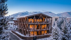 A modern building with a wooden facade and multiple windows, surrounded by snow-covered trees and mountains. - Mountain Residence CLARA (Brunico)
