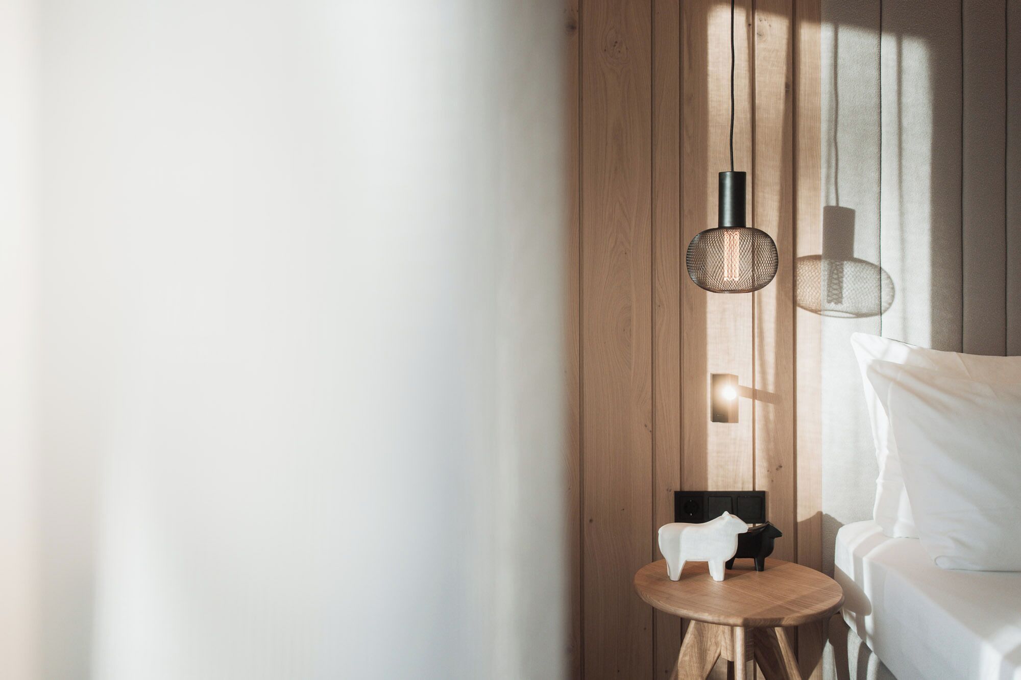 Un dormitorio moderno con una pared de madera, una mesa redonda de madera con una figurita de gato blanco, una silla negra y una lámpara colgante.