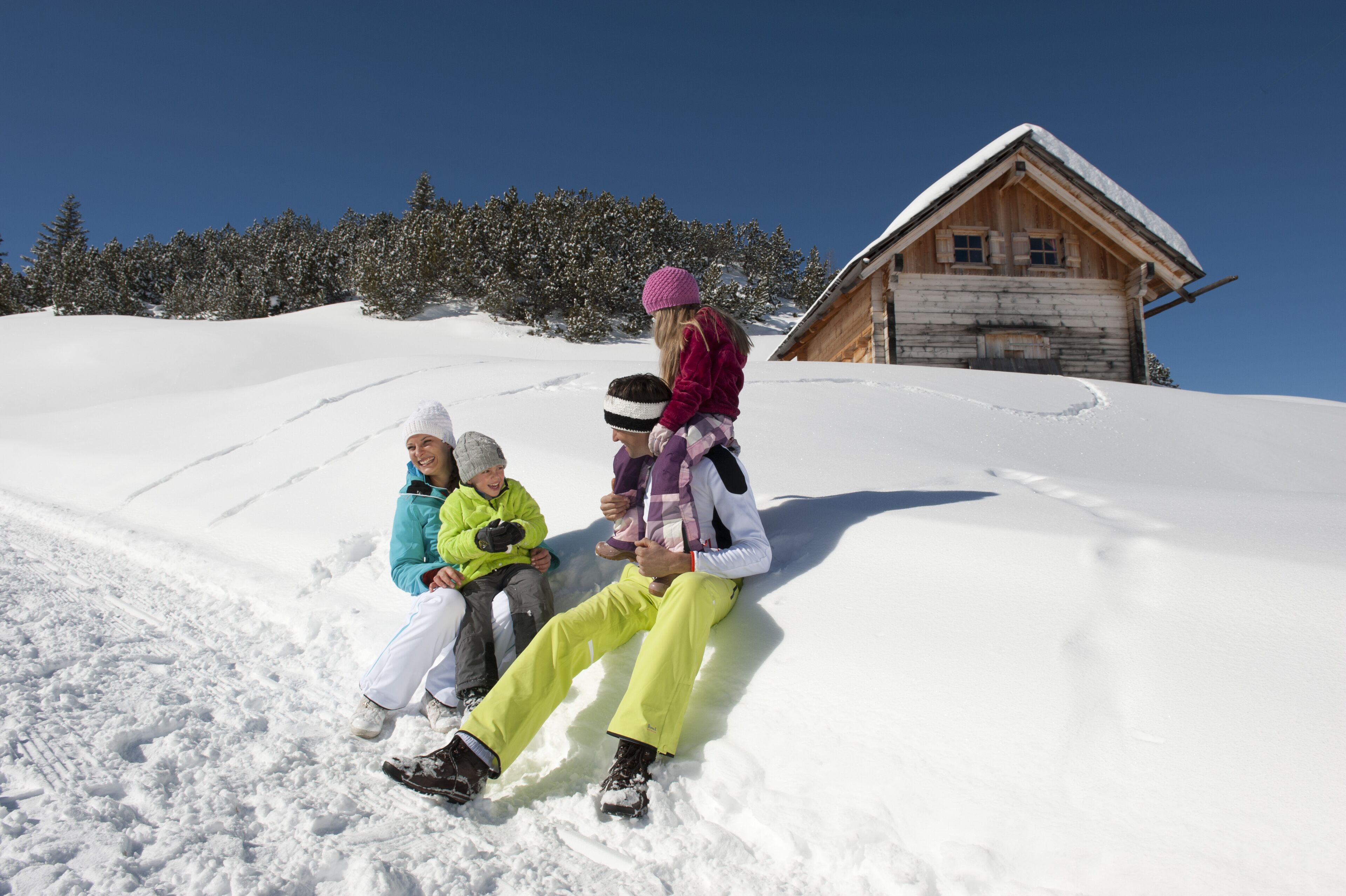 Eine Gruppe von Menschen sitzt auf einem mit Schnee bedeckten Hang, im Hintergrund ein Holzhaus.