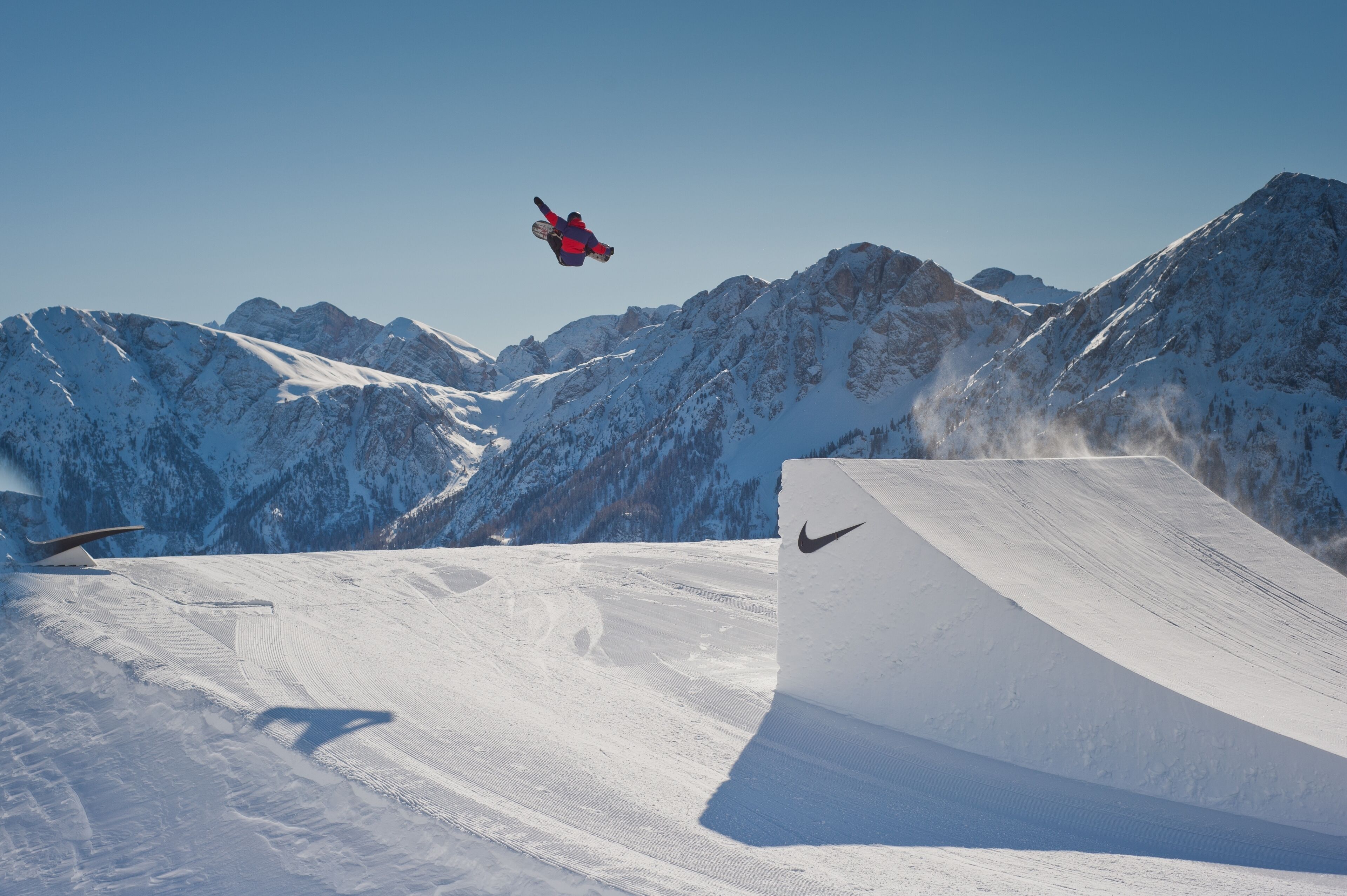 Un snowboarder en pleno vuelo realizando un truco en una ladera nevada de montaña, con un cielo despejado y montañas cubiertas de nieve al fondo.