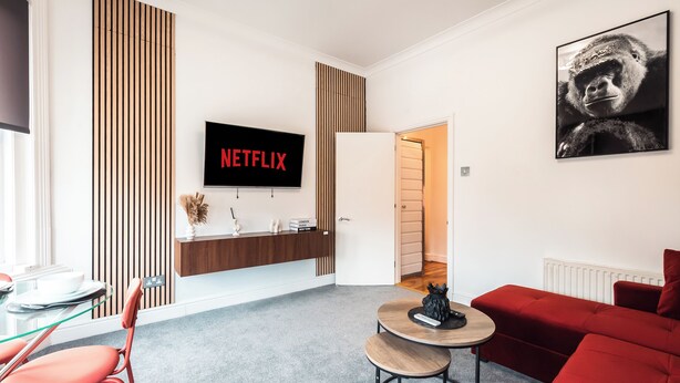 Un soggiorno moderno con un divano rosso, un tavolino piccolo e rotondo, un tavolino in vetro, una TV montata a parete e una porta che dà su un'altra stanza.