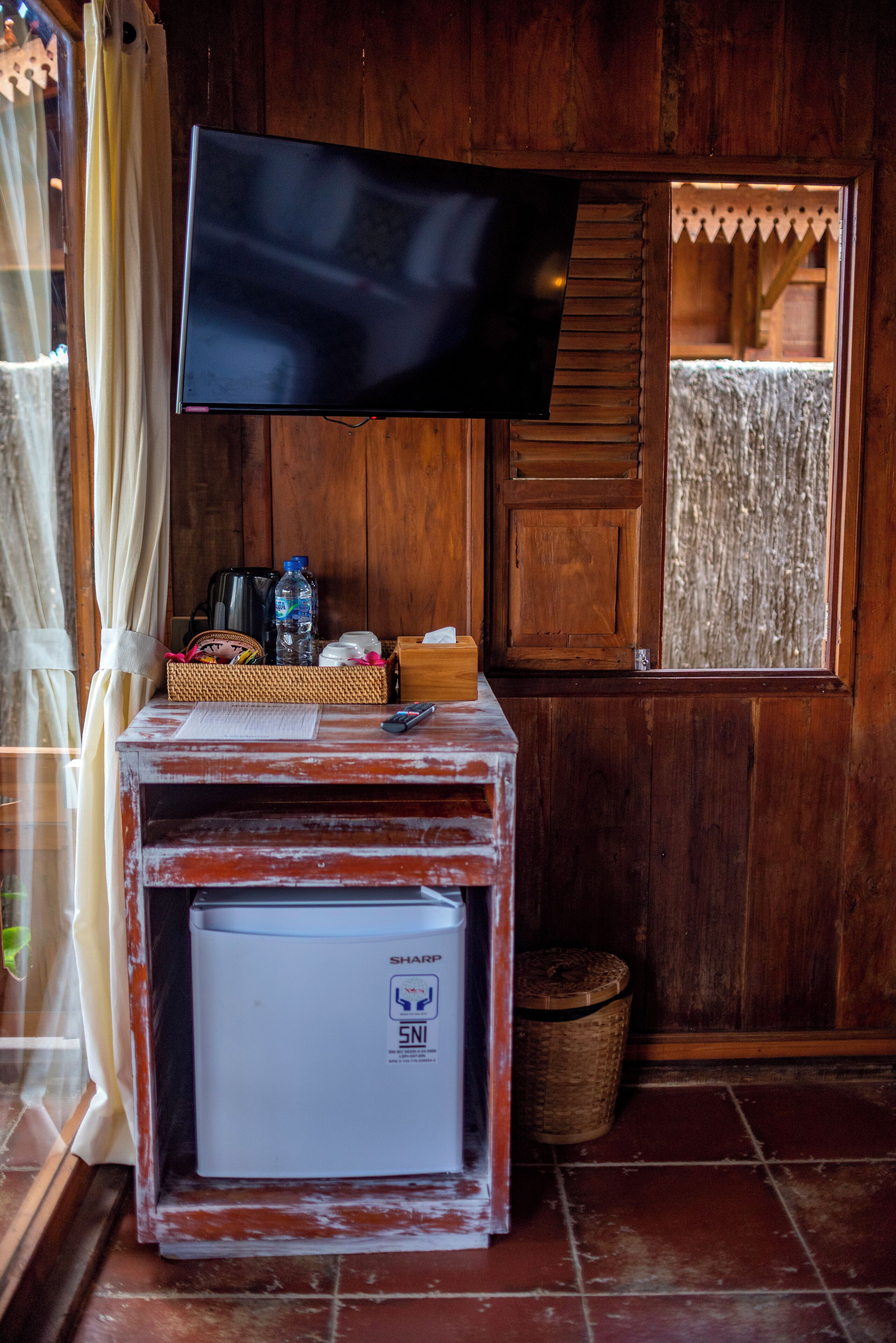 A wooden room with a television mounted on a wall, a small refrigerator, a basket with items, and a window with a curtain.