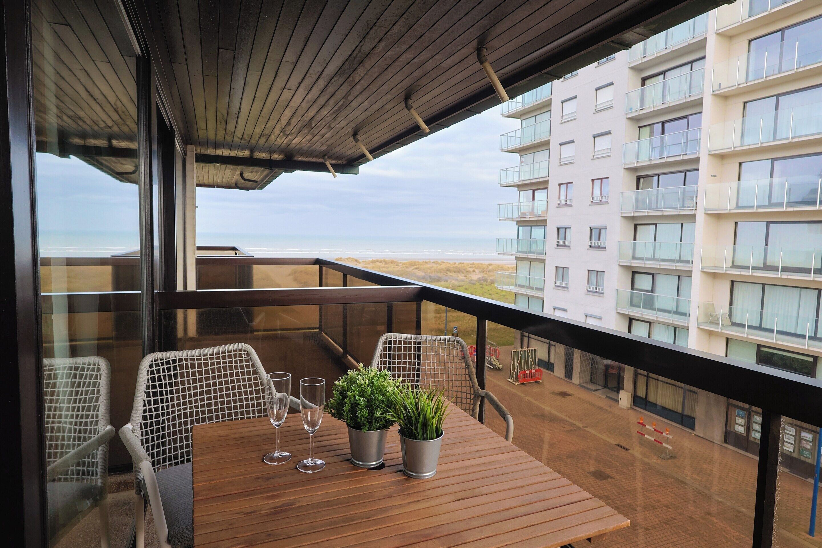 A balcony with a wooden table, two chairs, and two potted plants.