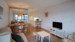 A modern living room with a dining area, a flat-screen TV on a white stand, and a kitchen in the background. - Studio by the Beach With Sea View (Koksijde)