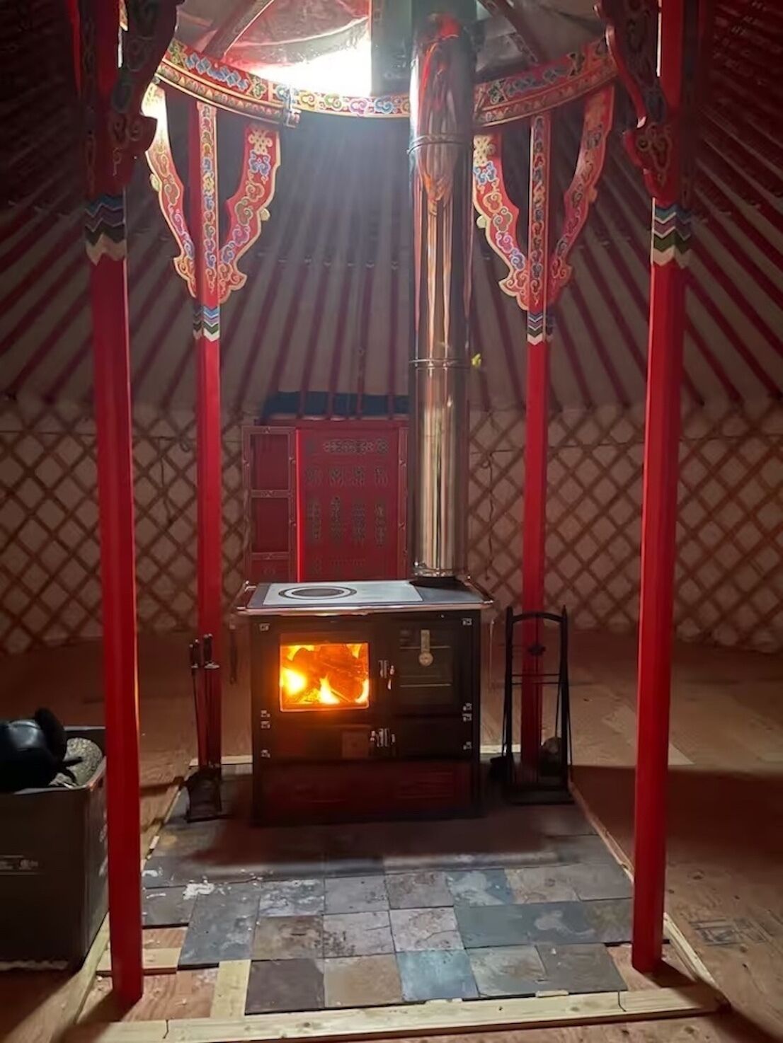 A traditional yurt with a wood-burning stove, red decorative pillars, and a tiled floor.