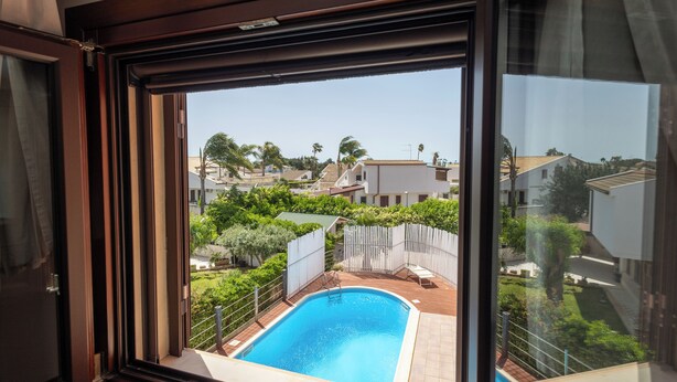A view from a window of a swimming pool surrounded by greenery and residential buildings. - Villa Stephanie with pool (Marina di Ragusa)