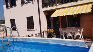 A swimming pool with a metal ladder, a table with chairs, and a yellow-striped awning over a building. - PO LAZISE HOUSE (Lazise)