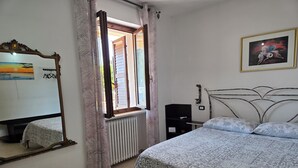 A bedroom with a bed, a chair, a radiator, a window with curtains, a wall-mounted shelf, and a framed picture. - PO LAZISE HOUSE (Lazise)
