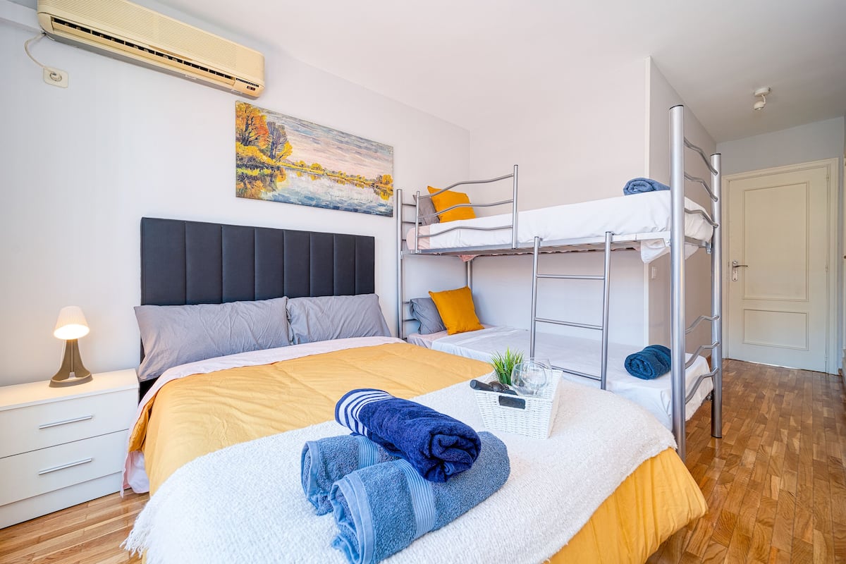A well-lit room with a bed, a nightstand, a wall-mounted air conditioner, and a bunk bed.