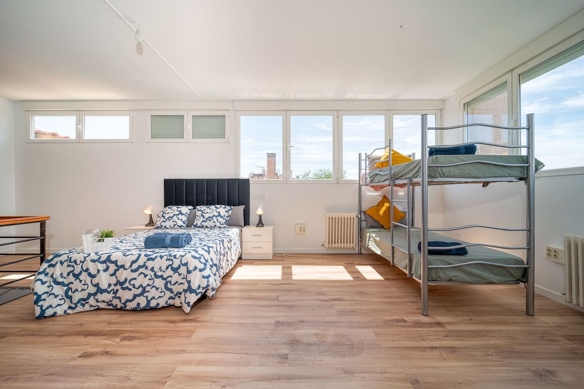 A modern bedroom with a bunk bed, a single bed with patterned bedding, a bedside table with a lamp, and a view of the city through large windows.