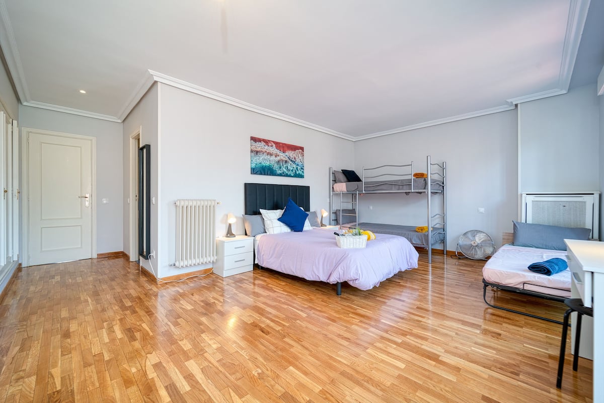 A bedroom with a wooden floor, a bed with a headboard, a bedside table, a radiator, a wall-mounted fan, and a bunk bed.