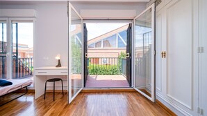 Une chambre moderne avec un plancher en bois, un bureau avec une chaise, un lit et une porte-fenêtre coulissante donnant sur un balcon avec vue sur les bâtiments et la verdure.